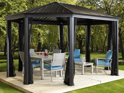 Ein Gartenpavillon mit schwarzem Metallgestell und einem festen Dach, der auf einer Holzplattform im Grünen steht, unter dem Pavillon stehen ein Gartentisch und Stühle