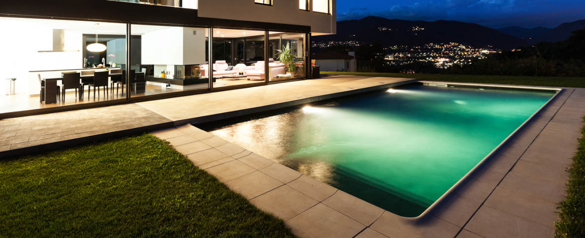 Beleuchteter moderner Außenpool bei Nacht vor einem Wohnhaus mit großen Glasfronten. Im Hintergrund sind Berge und eine Stadt mit Lichtern zu sehen.