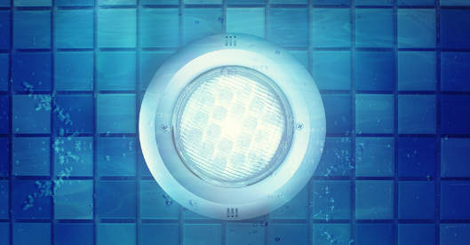 Eine runde Unterwasserlampe leuchtet an der gefliesten Wand eines Pools mit blauen Mosaikfliesen. Luftblasen steigen neben der Lampe auf.