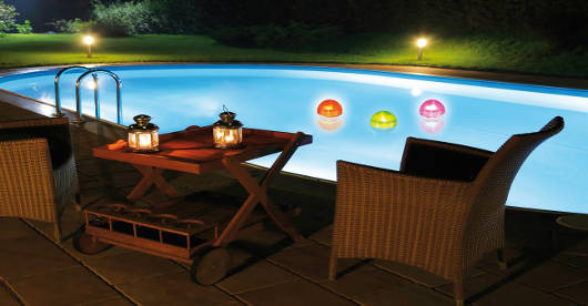 Ein beleuchteter Außenpool bei Nacht mit bunten Wasserbällen. Im Vordergrund stehen zwei Korbstühle und ein Teewagen mit Laternen auf einer Terrasse.