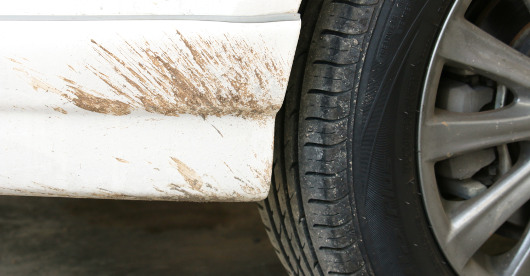 Ein Autoreifen neben einem verschmutzten weißen Kotflügel ist aus der Nähe zu sehen. Auf dem Lack befinden sich schlammige Kratzspuren, das Reifenprofil wirkt wenig abgefahren