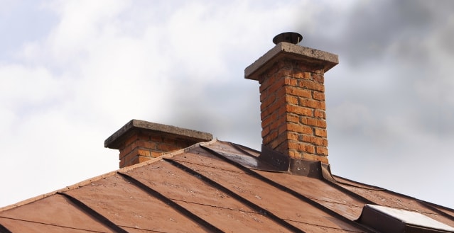 Zwei gemauerte Schornsteine mit Abdeckung ragen aus einem geneigten Metalldach in den wolkigen Himmel