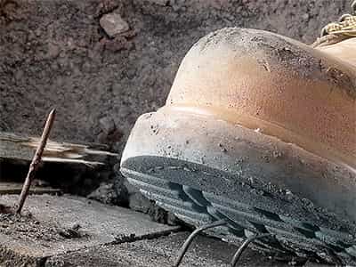 Ein Arbeitsschuh steht kurz davor, auf herausragende Nägel und einen rostigen Nagel zu treten. Die Szene zeigt eine potenzielle Verletzungsgefahr auf einer Baustelle.
