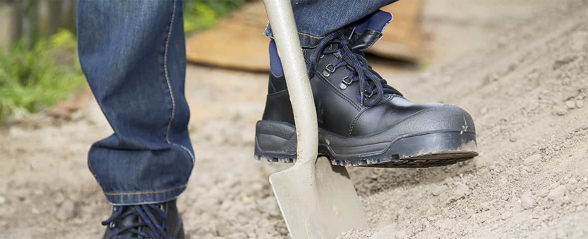 Eine Person mit Arbeitsschuhen tritt auf eine Schaufel, um Erde oder Sand zu bewegen. Die Szene zeigt einen Ausschnitt der Beine und des Werkzeugs.