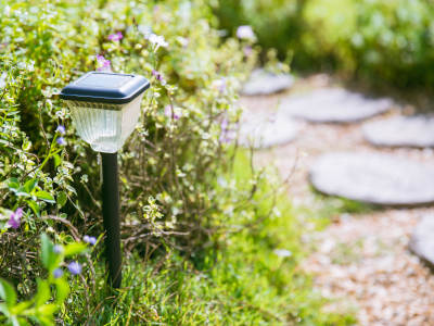 Eine kleine Solarlampe mit schwarzem Stab steht am Rand eines blühenden Beetes neben einem Gartenweg mit Trittsteinen. Die Lampe lädt sich tagsüber im Sonnenlicht auf