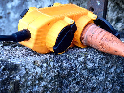 Eine orange-gelbe wetterfeste Outdoor-Steckdose liegt auf einem rauen Betonblock. Zwei schwarze Kabel sind eingesteckt