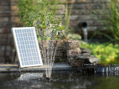 Ein Gartenteich mit kleiner Springbrunnenfontäne, links am Beckenrand steht ein schräg aufgestelltes Solarpanel, das Sonnenlicht einfängt