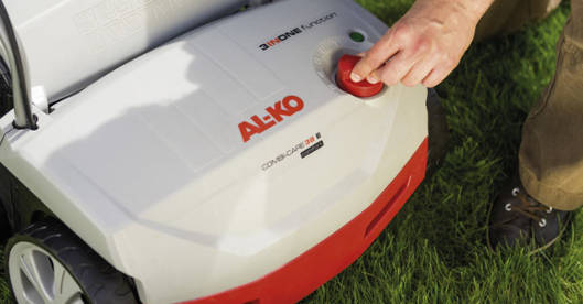 Eine Person stellt an einem elektrischen Vertikutierer auf einer Rasenfläche die Arbeitstiefe ein