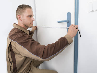 Foto von einem Mann in brauner Arbeitskleidung, der mit einem Maßband die Breite eines Türblatts misst, die Tür ist weiß mit blauer Klinke